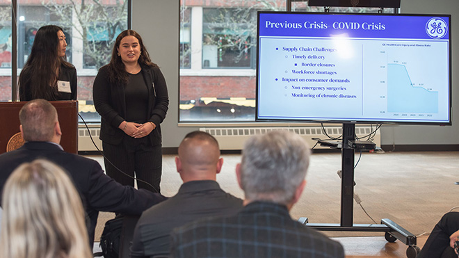 Two MBA students stand at a podium next to a screen that reads "Previous Crisis- COVID Crisis"