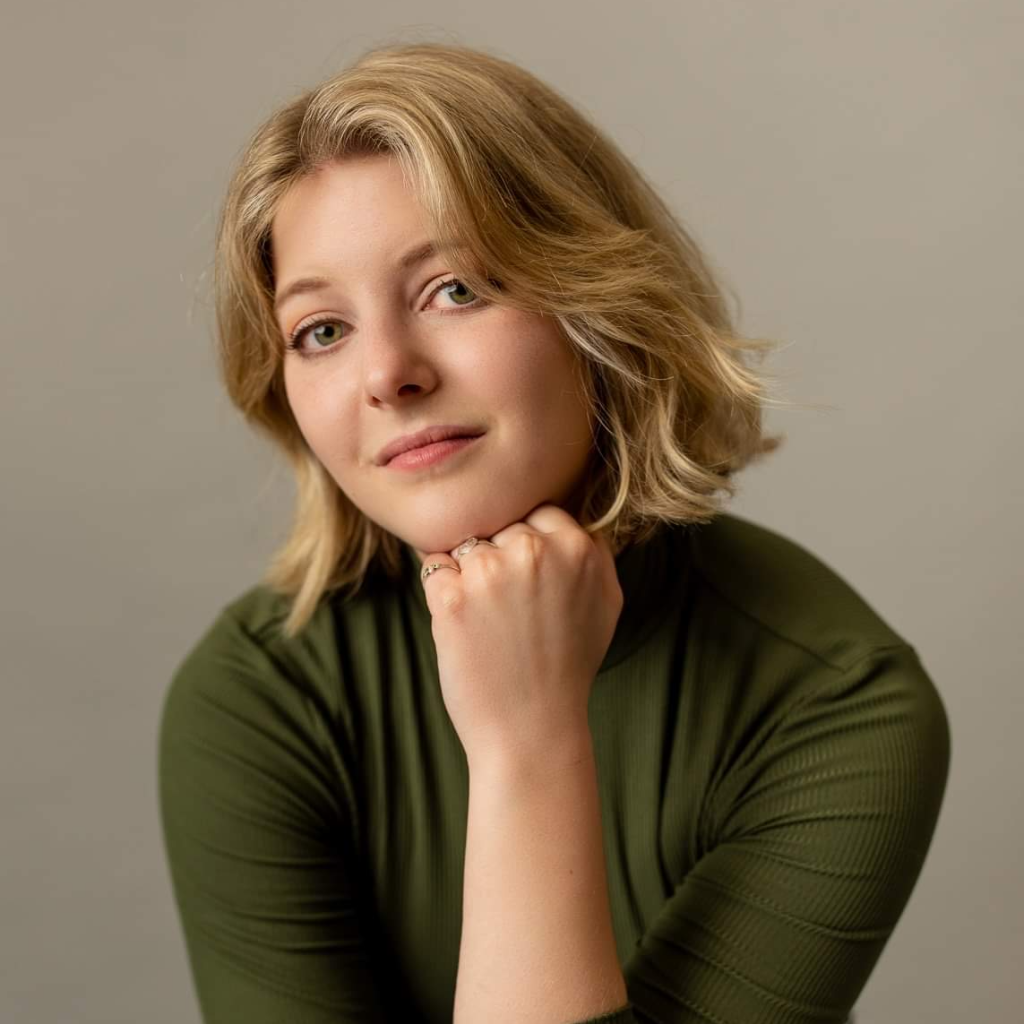 Brooke Aubin. Brooke is a white, blonde person with shoulder length hair wearing a green shirt. Brooke is resting their chin on one hand and smiling slightly.