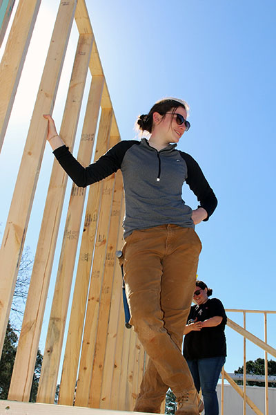 Genevieve standing with her hand on stud of a framed wall outside on a sunny day wearing sunglasses