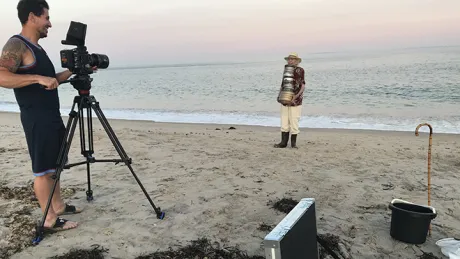 A director with camera films an actor holding film reels on a beach