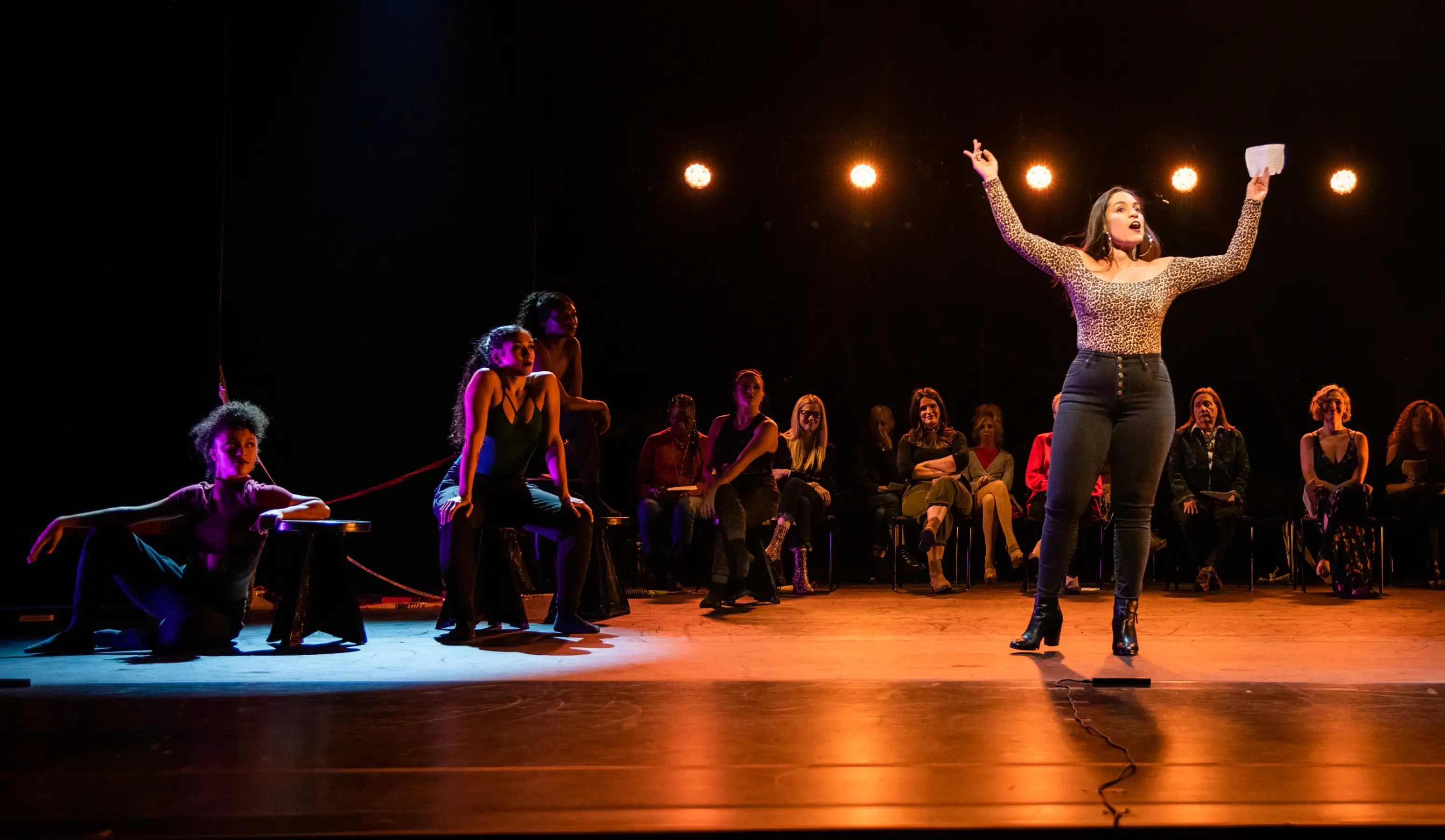 Performer sings on stage with raised arms as dancers pose and audience members watch under dramatic lighting.