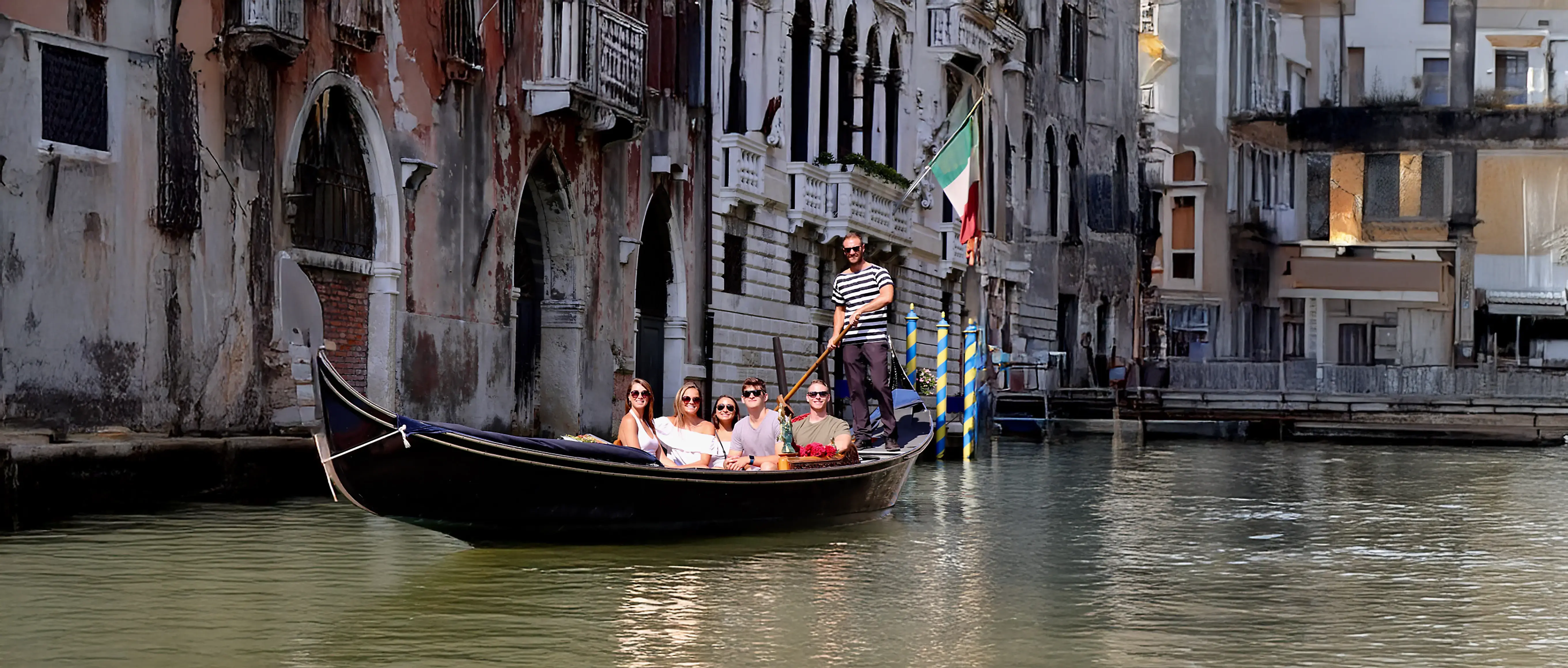 Students on a gondola ride through a canal in Venice, Italy, with historic buildings and an Italian flag in the background.