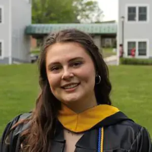 Roger Williams University graduate in cap and gown smiles outdoors on campus.