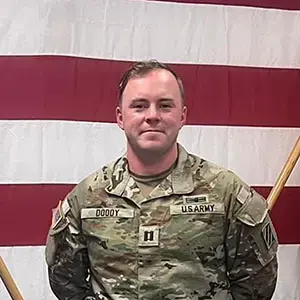 Student in U.S. Army uniform stands in front of an American flag.